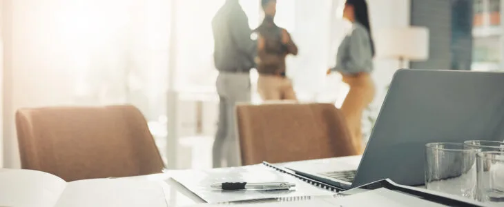 Office setting with a laptop, notepad, and pen in foreground, blurred figures discussing in background, symbolizing collaboration and startup intellectual property protection.
